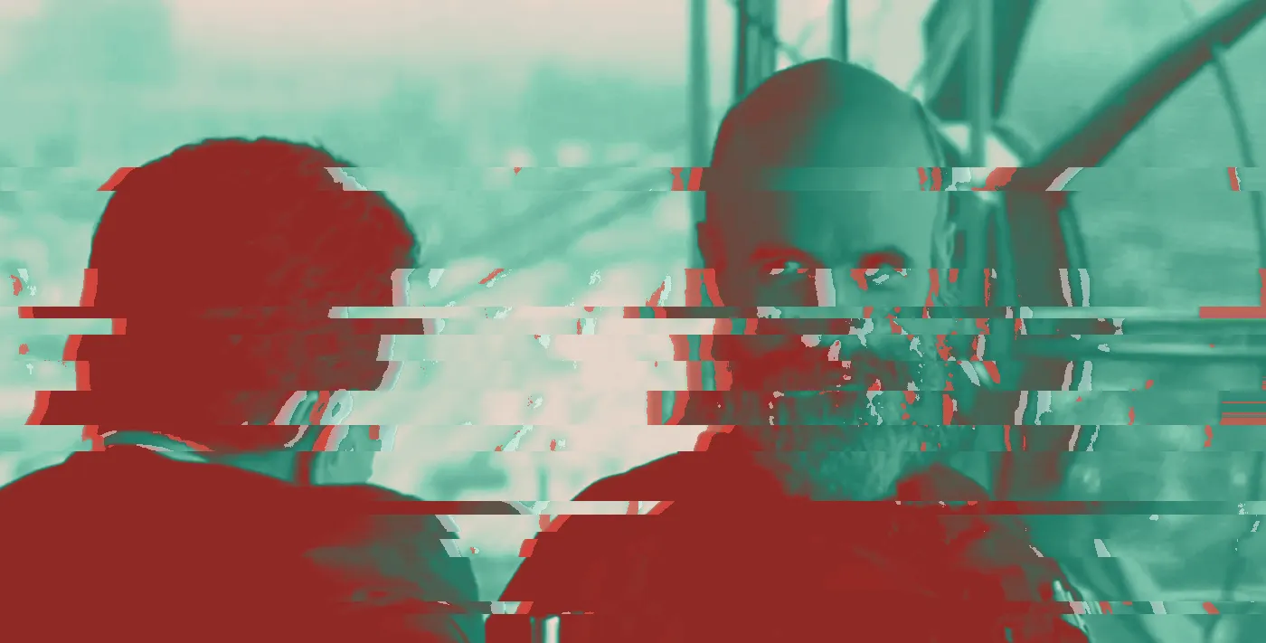 Film still of a bald, grey-bearded man talking to a man seen from behind