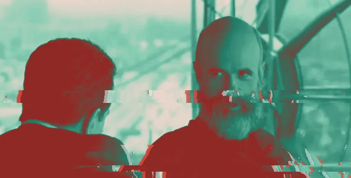Film still of a bald, grey-bearded man talking to a man seen from behind